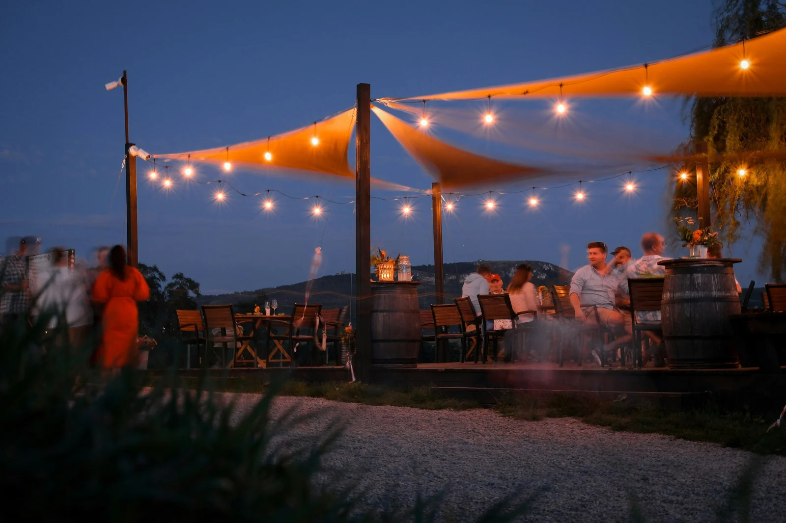 Outdoor restaurant patio at dusk with string lights, barrel tables, and guests gathered under a canvas shade sail