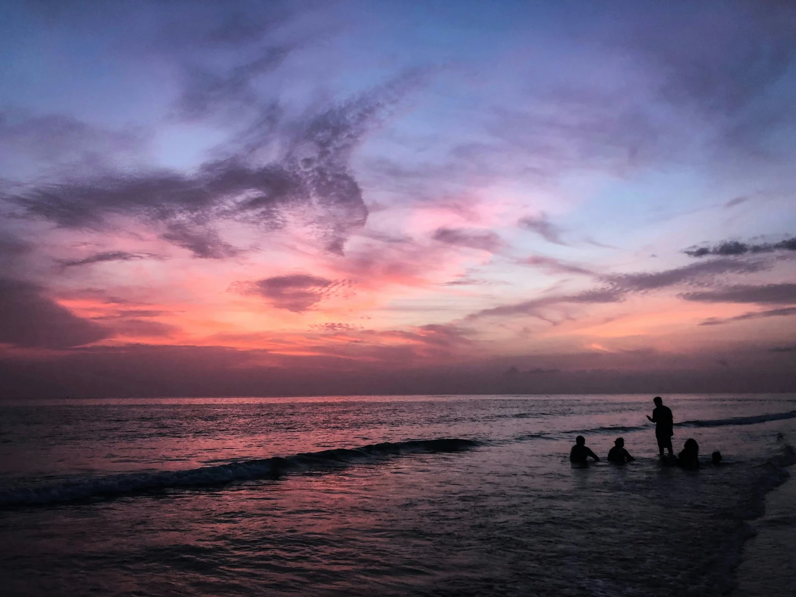 Silhouettes of people wading at the Gulf Coast shoreline during a vivid purple and pink sunset