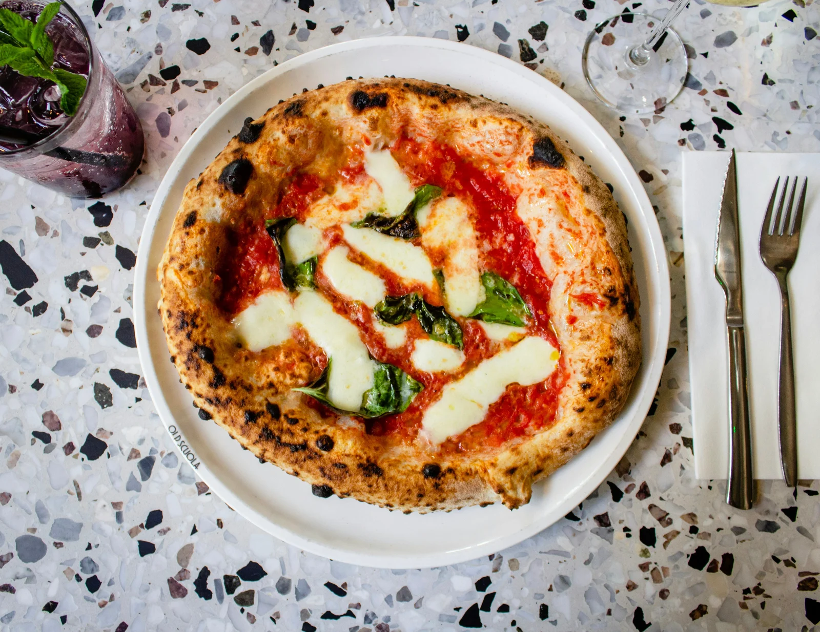 A Neapolitan margherita pizza on a white plate at a restaurant table, with fresh mozzarella, basil, and a charred crust