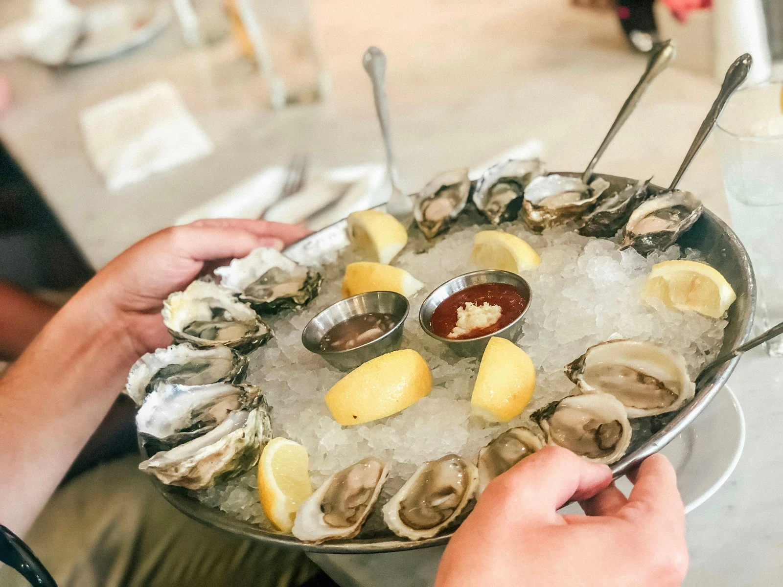 A platter of fresh oysters on ice with lemon wedges and mignonette, being carried to a table