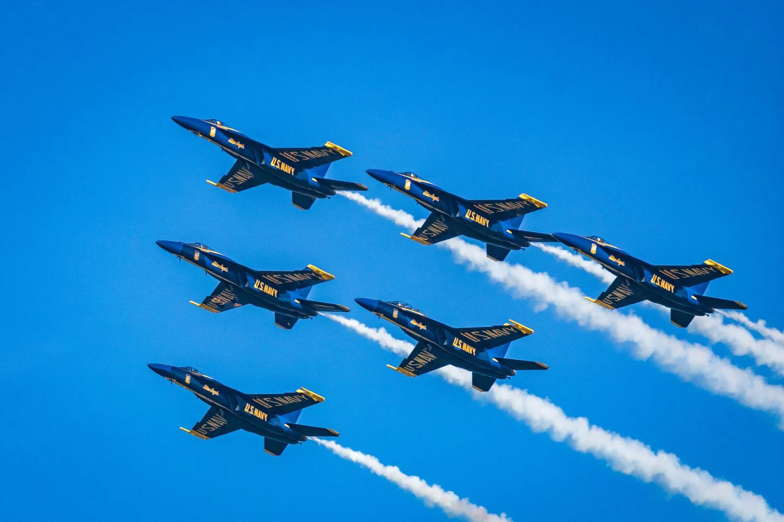Six U.S. Navy Blue Angels jets flying in tight diamond formation with white smoke trails against a deep blue sky